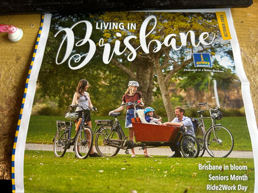 Photo of the front cover of "Living In Brisbane" magazine, the photo on the front shows several bikes, including a cargo bike carrying children, next to a bike path in a grassy park with shady trees in the background. Writing in the bottom right suggests an article on Ride2Work Day is inside.
